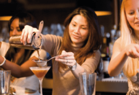 Un grupo de participantes en una clase de coctelería, reunidos alrededor de una estación de bar, mezclando ingredientes y preparando bebidas bajo la guía de un instructor.