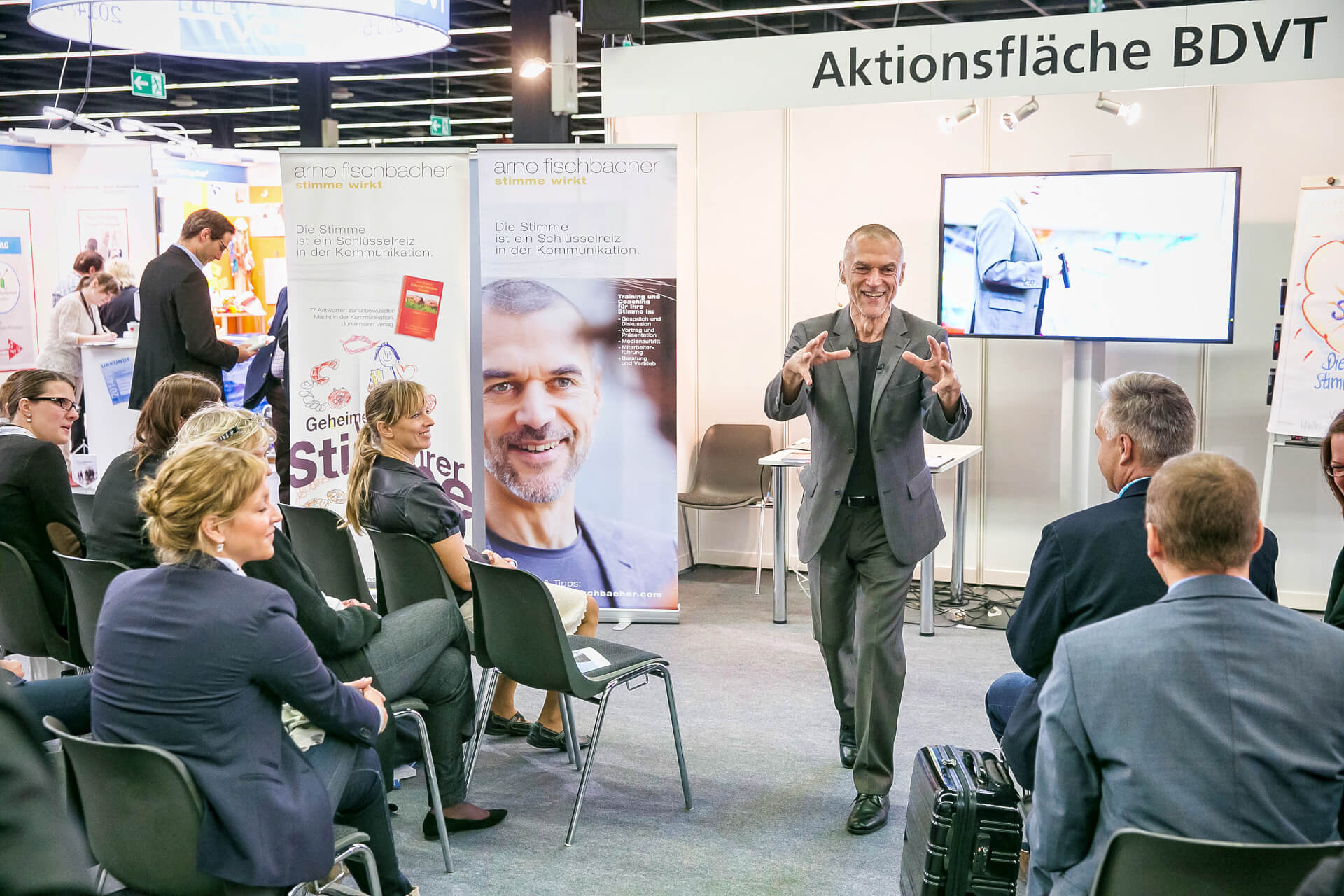 Un orador da una charla dinámica en una conferencia frente a una audiencia atenta; Al fondo, se ven pancartas publicitarias y una pantalla de televisión.