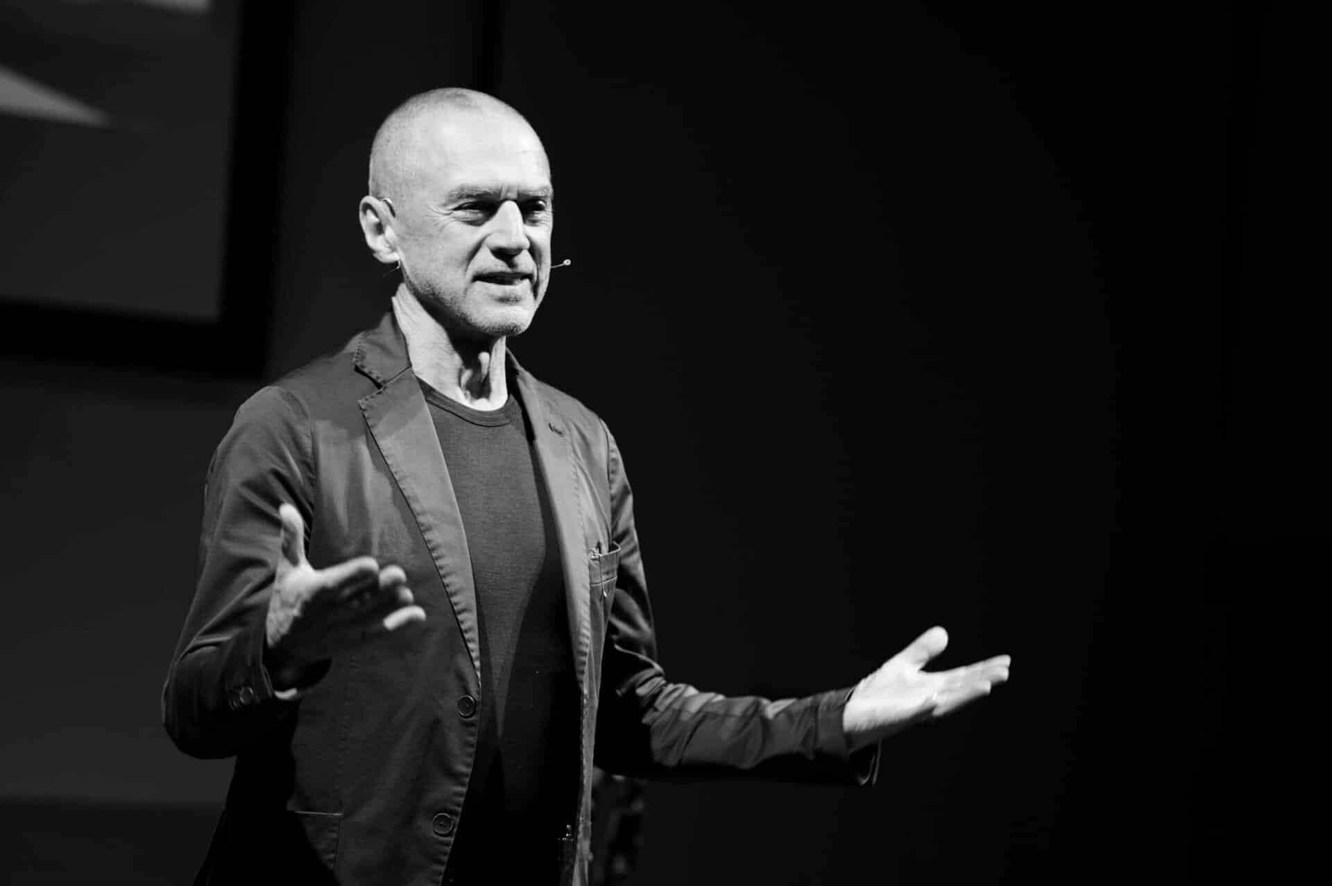 22-04-28 Freilassing Lokwelt 3 Un hombre con un blazer negro y una camiseta se para en un escenario y habla con las manos abiertas durante la presentación de información importante para la prensa. El fondo es oscuro y la luz se dirige hacia él.