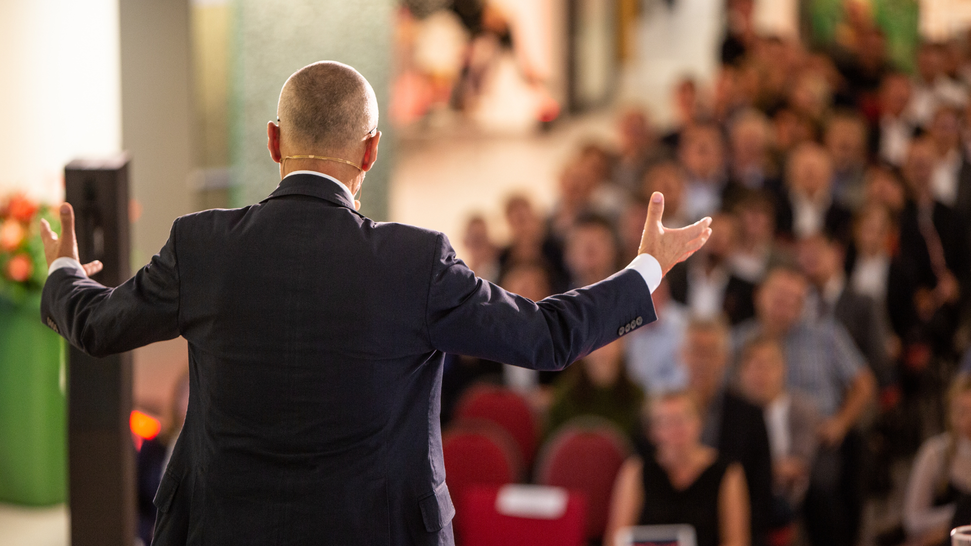 Un orador con un micrófono de diadema habla a una audiencia en una sala grande y hace un gesto con los brazos abiertos sobre "Más éxito en el liderazgo". El público está formado por hombres y mujeres y es un poco borroso.