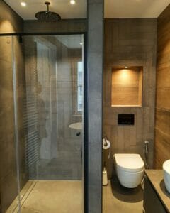 Modernes Badezimmer mit einer Glasdusche, die am Gehäuse befestigt ist, einer Wandtoilette zur Reduzierung und einem effektiven Regal über der Toilette. Die Wände sind mit grauen und holzähnlichen Oberflächen gefliest.