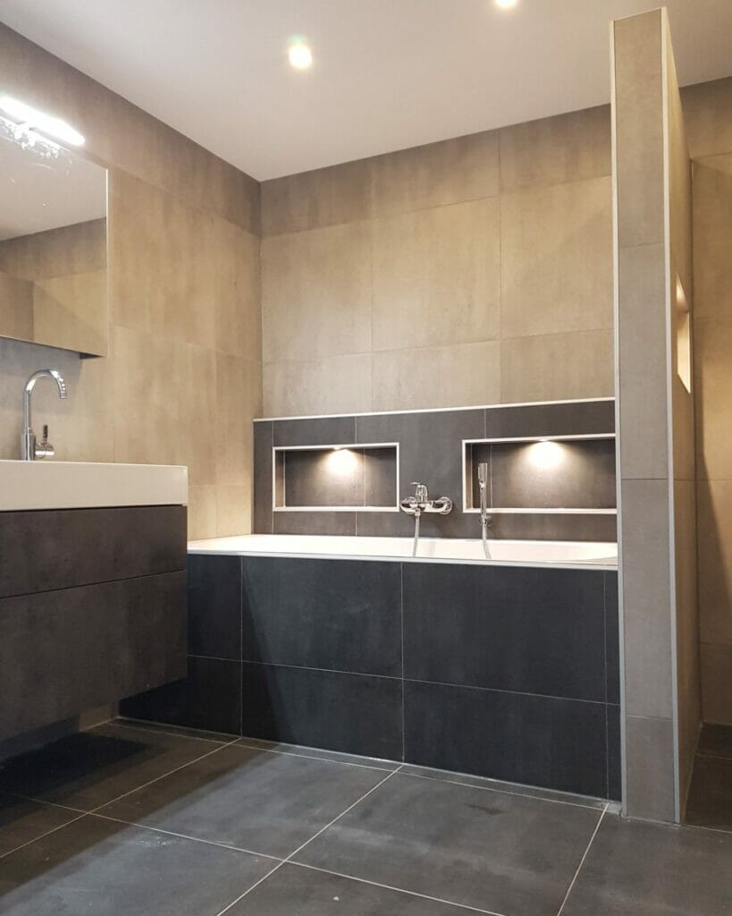 Modernes Badezimmer mit eingebauter Badewanne, Einbaubeleuchtung, minimalistischem Waschbecken und großen grauen Fliesen an Wänden und Boden, ausgestattet mit innovativen Beleuchtungslösungen für das Badezimmer.