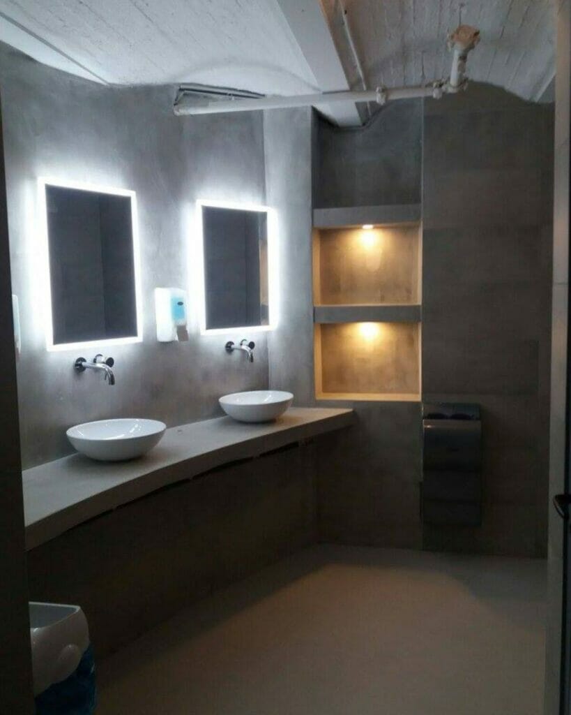 Ein modernes Badezimmer hat zwei Waschbecken mit beleuchteten Spiegeln oben und eingebaute Regale mit Strahlern auf der rechten Seite. Der Raum präsentiert sich in einem minimalistischen Design aus grauem Beton, das durch innovative Beleuchtungslösungen für das Badezimmer ergänzt wird.