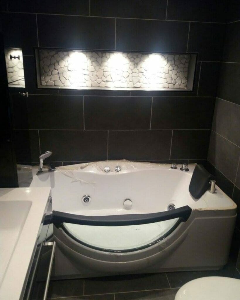 Ein modernes Badezimmer mit dunkel gefliesten Wänden verfügt über eine weiße Whirlpool-Badewanne, eine Kieselstein-Akzentwand und innovative Beleuchtungslösungen für das Badezimmer, die das nahe gelegene Waschbecken mit reflektierendem Spritzschutz ergänzen.