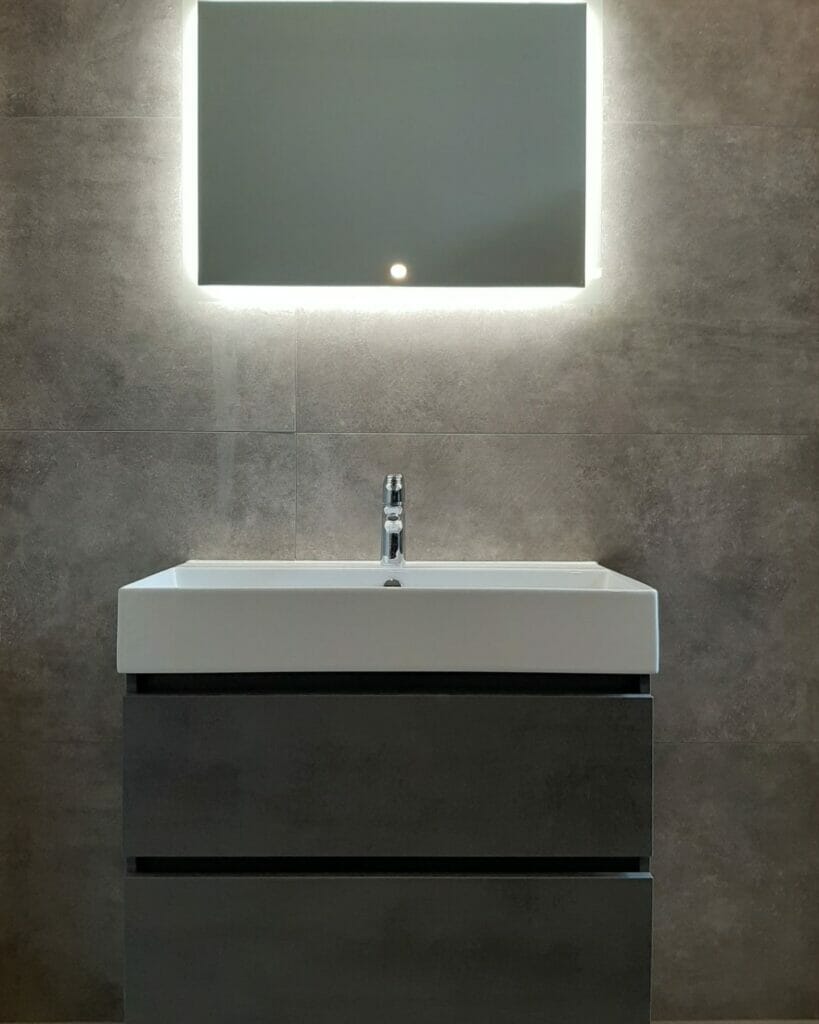 Modernes Waschbecken mit einem rechteckigen Spiegel darüber, der von einer hintergrundbeleuchteten LED beleuchtet wird. Die Wand ist mit großen, grauen Fliesen verkleidet – ein ideales Beispiel für Beleuchtungslösungen im Badezimmer.