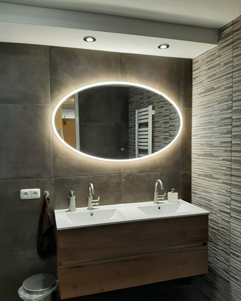Ein modernes Badezimmer mit einem Doppelwaschbecken, einem ovalen beleuchteten Spiegel, grau gefliesten Wänden und einem Handtuch, das neben einer Steckdose hängt. Deckeneinbauleuchten bieten brillante Beleuchtungslösungen für das Badezimmer, um den Raum zu beleuchten.