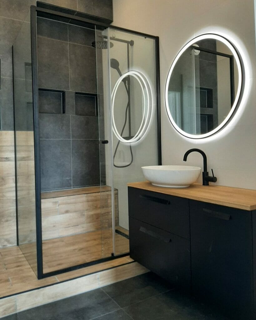 Modernes Badezimmer mit Duschkabine aus Glas, rundem beleuchtetem Spiegel, Waschbecken in Schwarz und Holz, Waschbecken, schwarzen Armaturen und dunklen Fliesenwänden. Durchdachte Beleuchtungslösungen für das Badezimmer unterstreichen die schlanke Ästhetik des Raums.