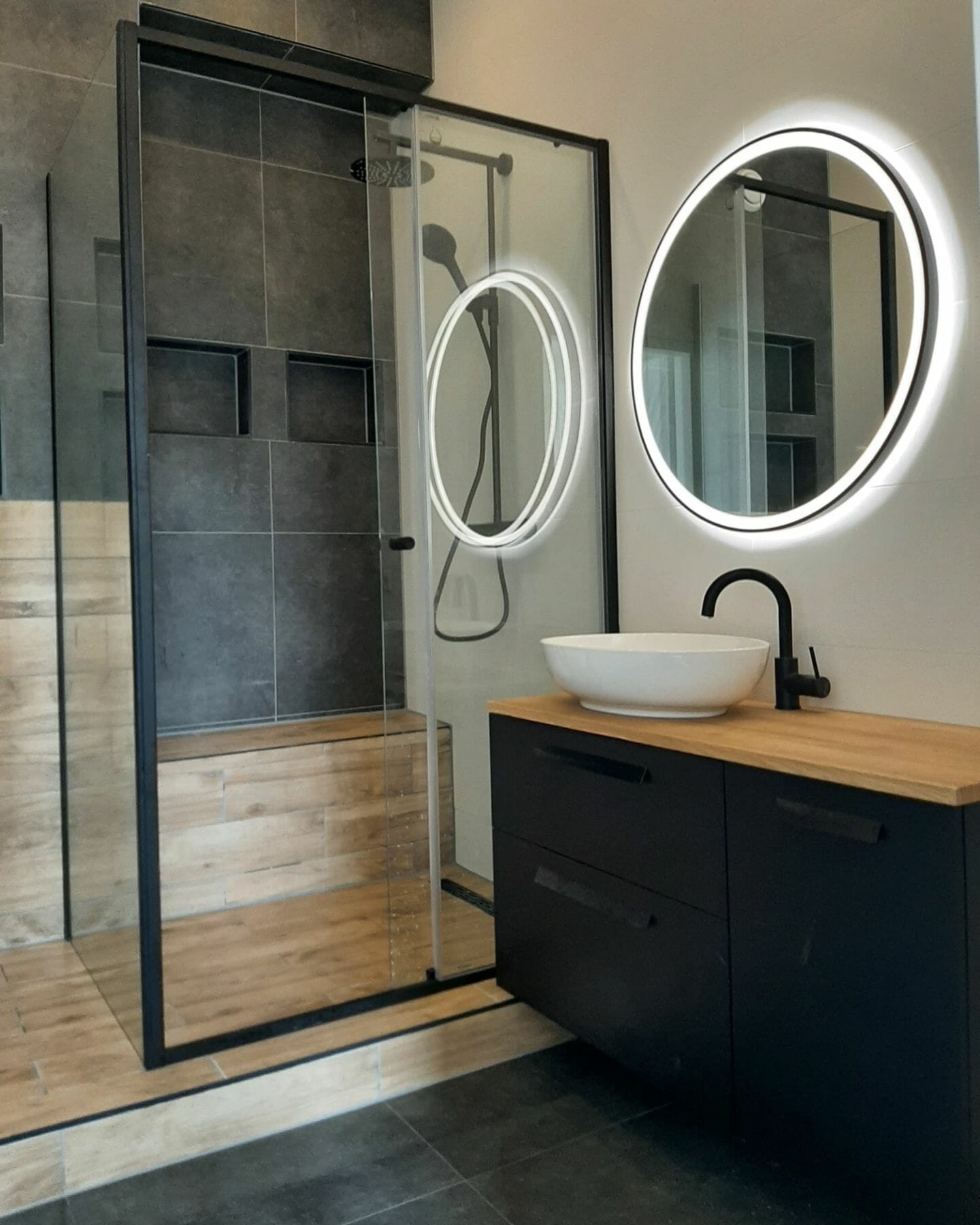 Modernes Badezimmer mit Duschkabine aus Glas, rundem beleuchtetem Spiegel, Waschbecken in Schwarz und Holz, Waschbecken, schwarzen Armaturen und dunklen Fliesenwänden. Durchdachte Beleuchtungslösungen für das Badezimmer unterstreichen die schlanke Ästhetik des Raums.