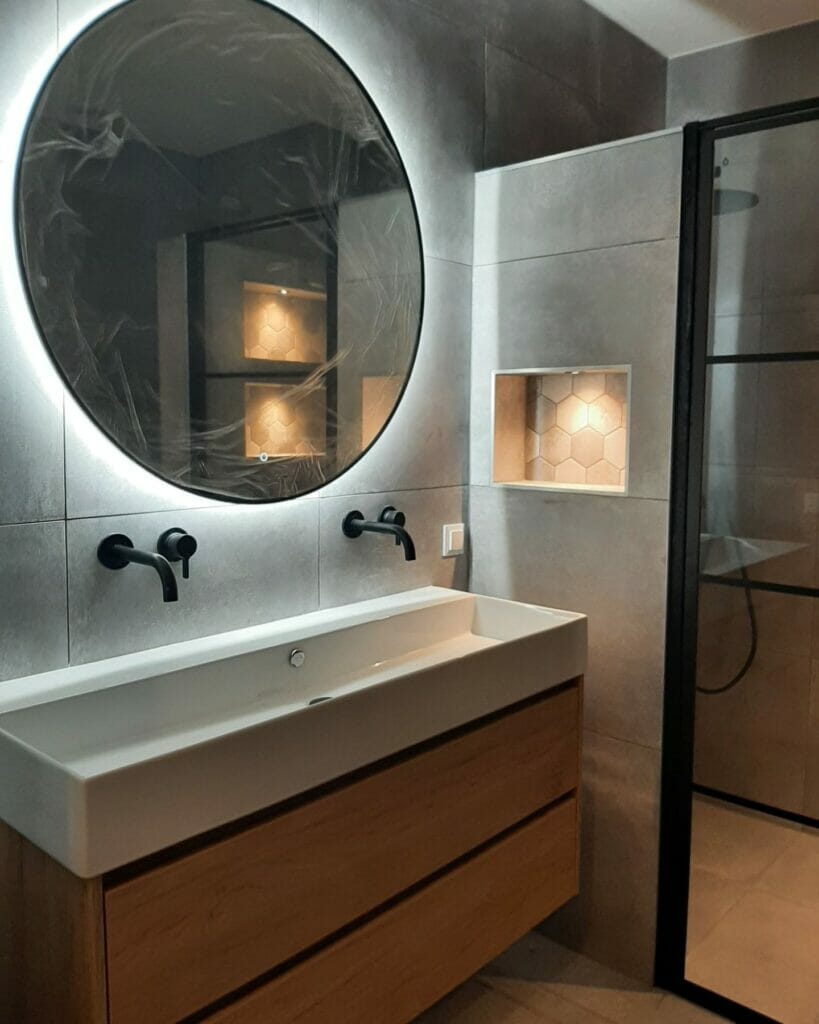 Modernes Badezimmer mit einem großen runden Spiegel mit Beleuchtung über einem Waschtisch mit zwei Waschbecken, schwarzen Armaturen, grauen Fliesenwänden und einer Duschkabine aus Glas, ausgestattet mit innovativen Beleuchtungslösungen für das Badezimmer.