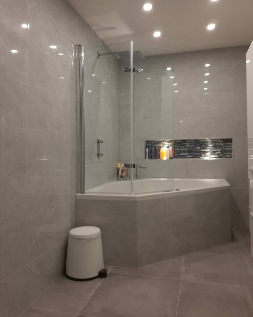 Ein modernes Badezimmer mit Badewannen-Dusch-Kombination, Glastrennwand und grau gefliesten Wänden und Fußboden. Ein weißer Hocker steht neben der Badewanne unter stilvollen Badezimmerbeleuchtungslösungen. Toilettenflaschen sind auf einem Regal im Duschraum sichtbar.