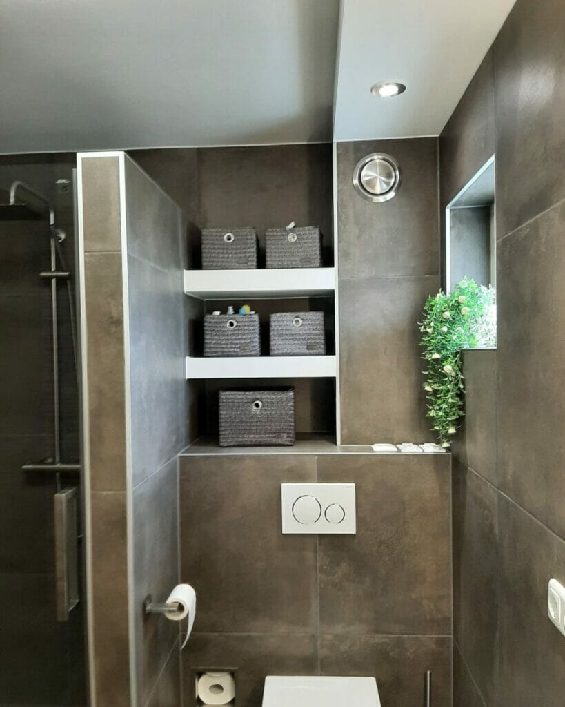 Modernes Badezimmer mit grauen Fliesen, einem wandhängenden Kleiderschrank, Badezimmerbeleuchtungslösungen mit Regalen mit geflochtenen Körben, einem kleinen Fenster mit einer Pflanze und einem Duschbad aus Glas. Toilettenpapier wird an der Wand befestigt.