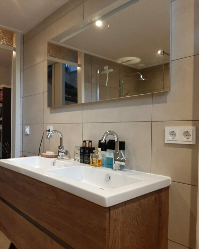 Modernes Waschbecken im Badezimmer mit großem Spiegel, Doppelarmaturen, einem Holzwaschbecken und verschiedenen Toilettenartikeln auf der Arbeitsplatte. An der gefliesten Wand befinden sich zwei Steckdosen, die durch effiziente Beleuchtungslösungen für das Bad ergänzt werden.
