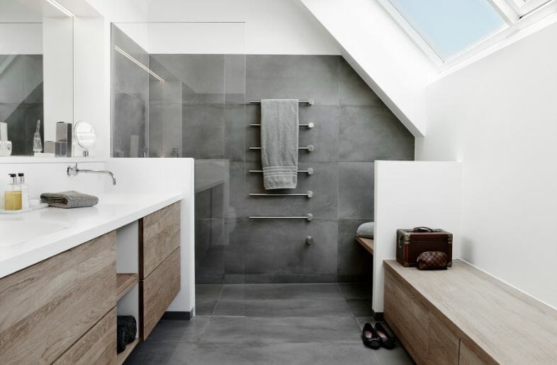 Modernes Badezimmer mit grauen Fliesen, begehbarer Dusche, Oberlicht, weißer Arbeitsplatte, Holzschränken und stilvollem Handtuchhalter, der auf Funktionalität ausgelegt ist.