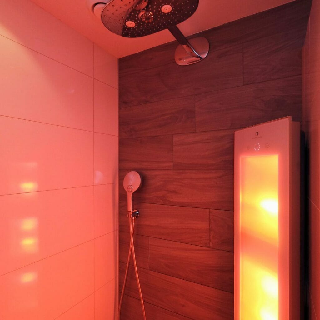 Eine Duschkabine mit dunklen Fliesen in Holzoptik an der Rückwand, ein Regenduschkopf und eine Handbrause sowie eine leuchtende Wandheizung, die rotes Licht abgibt – eine der besten Beleuchtungslösungen für das Badezimmer.