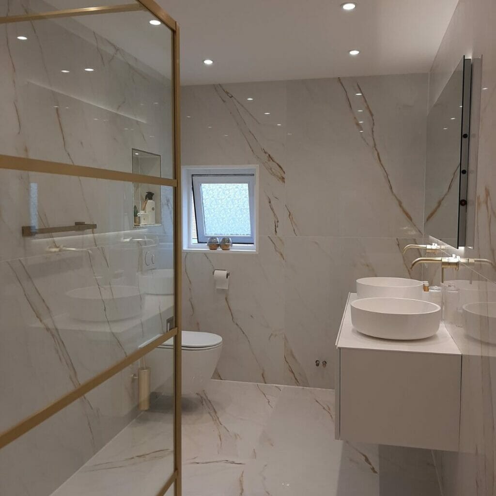 Ein modernes Badezimmer mit einer Glasdusche, weißen Waschbecken auf einem schwimmenden Waschbecken, einer Toilette und Beleuchtungslösungen für das Badezimmer. Wände und Boden sind mit weißem Marmor mit goldenen Akzenten verkleidet, ergänzt durch ein kleines Fenster.