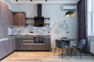 Moderne Küche mit Holzschränken, sechseckiger Fliesenrückwand, schwarzen Geräten, rundem schwarzem Esstisch mit vier grauen Stühlen und großem Fenster mit dunklen Vorhängen.