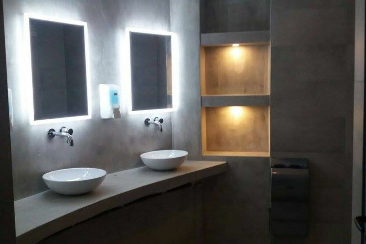 Ein modernes Badezimmer hat zwei Waschbecken mit beleuchteten Spiegeln oben und eingebaute Regale mit Strahlern auf der rechten Seite. Der Raum präsentiert sich in einem minimalistischen Design aus grauem Beton, das durch innovative Beleuchtungslösungen für das Badezimmer ergänzt wird.