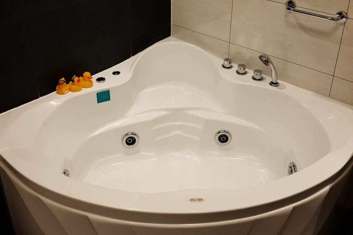 Weiße Eckbadewanne mit mehreren Düsen, silbernen Bedienelementen und drei Gummienten am Rand. Ein Metallgriff ist sicher an der gefliesten Wand befestigt, was dieses Badezimmer zu einem familienfreundlichen Badezimmer macht, das sich perfekt zum sicheren Entspannen mit der Familie eignet.