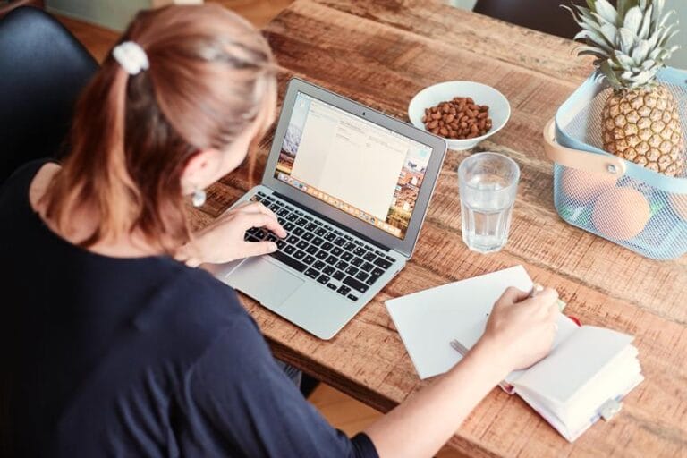 Person, die an einem Tisch sitzt und auf einem Laptop tippt, mit einem Notizbuch, einem Glas Wasser, einer Schüssel mit Nüssen und einem Korb mit einer Ananas und anderen Früchten in der Nähe; Erinnert an die gemütliche Atmosphäre, die oft in charmanten Küchengeschäften zu finden ist.