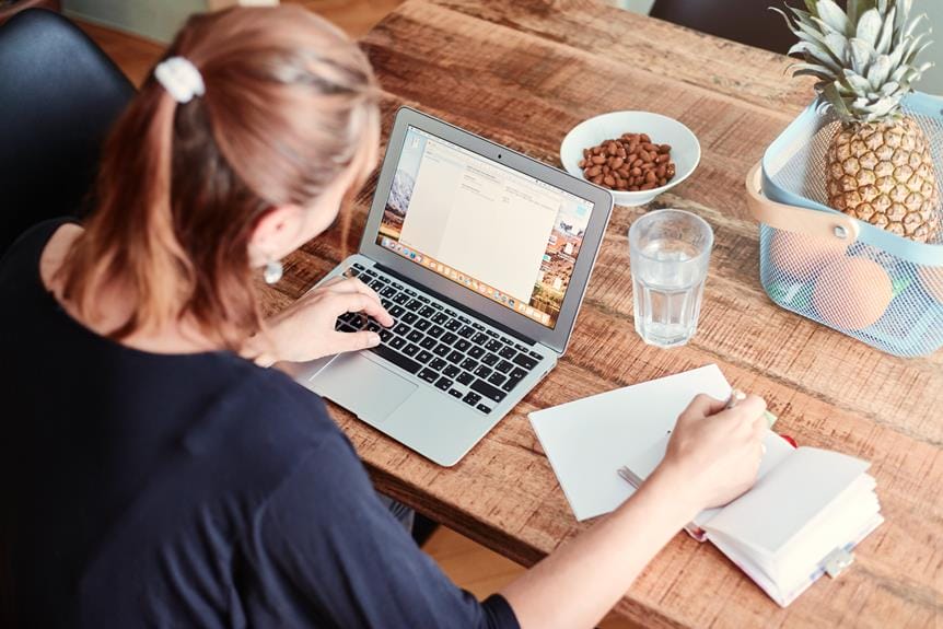 Person, die an einem Tisch sitzt und auf einem Laptop tippt, mit einem Notizbuch, einem Glas Wasser, einer Schüssel mit Nüssen und einem Korb mit einer Ananas und anderen Früchten in der Nähe; Erinnert an die gemütliche Atmosphäre, die oft in charmanten Küchengeschäften zu finden ist.