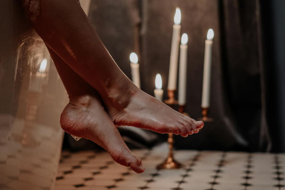 Nahaufnahme der Beine und Füße einer Person, die auf dem Rand einer Badewanne ruht. Auf dem Boden liegen Wassertropfen, umgeben von brennenden Kerzen, die an Auslagen erinnern, die man in einem Badezimmergeschäft findet. Das Zimmer verfügt über Fliesenböden und gedämpfte Beleuchtung.
