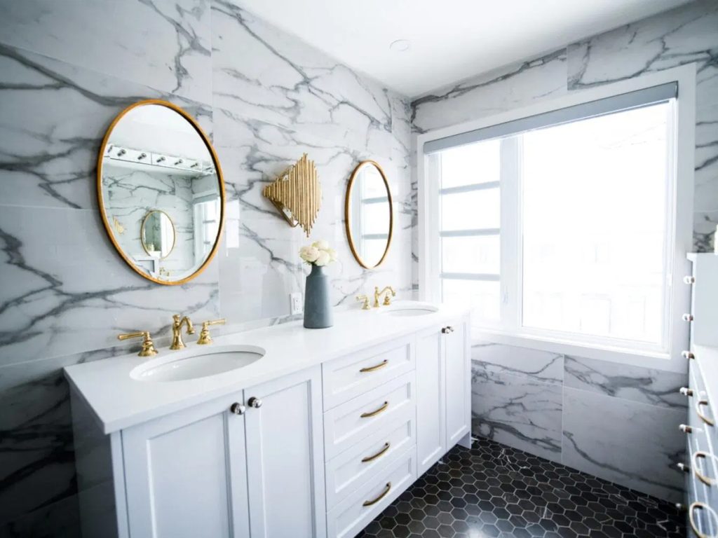 Erleben Sie ein modernes DIY-Badezimmer mit zwei runden Spiegeln, weißen Marmorwänden und zwei Waschbecken, die mit eleganten goldenen Armaturen verziert sind. Das zentrale Blumenarrangement ergänzt die sechseckigen Bodenfliesen und das große Fenster auf der rechten Seite und schafft einen einladenden Rückzugsort.