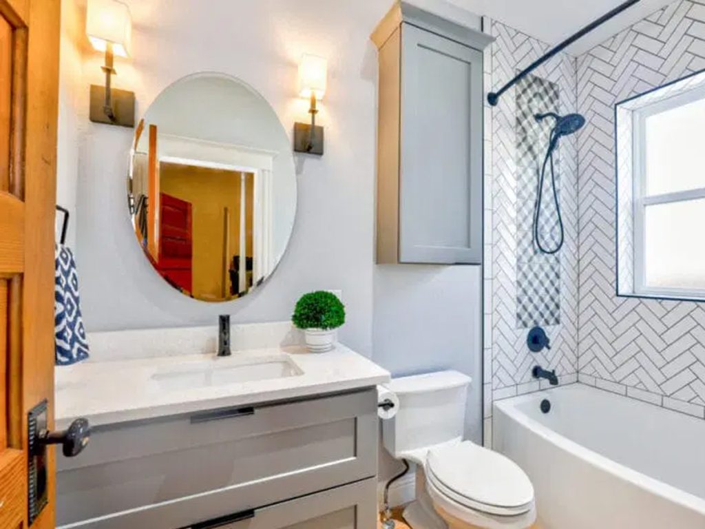 Zeitgenössische Badezimmerrenovierungen zeigen ein modernes Badezimmer mit einem grauen Waschbecken, einem ovalen Spiegel und Wandlampen. Es verfügt über eine Badewanne mit Fischgrätmuster und ein einladendes Fenster. Die Arbeitsplatte wird durch eine kleine Pflanze akzentuiert, die für einen Hauch von Natur sorgt.