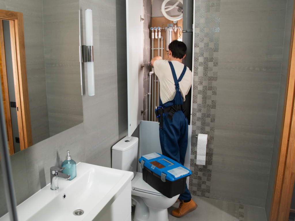 Een loodgieter in een blauwe overall pakt vakkundig een badkamerrenovatie aan, waarbij hij leidingen bij het toilet en de wastafel repareert, terwijl hij een gereedschapskist op de toiletdeksel legt.