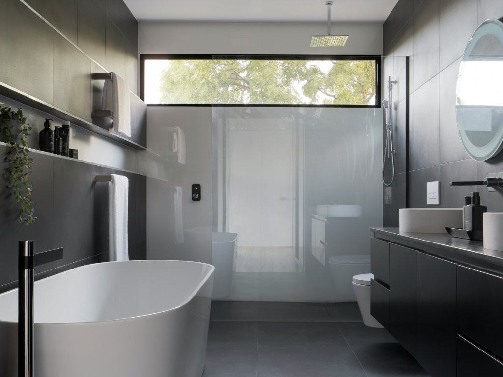 Modernes Badezimmer mit Badmöbeln mit freistehender Badewanne, schwimmendem Waschbecken, Wandschrank und großem Milchglasfenster. Dunkle Fliesen und ein runder Spiegel runden das schlichte Design ab.