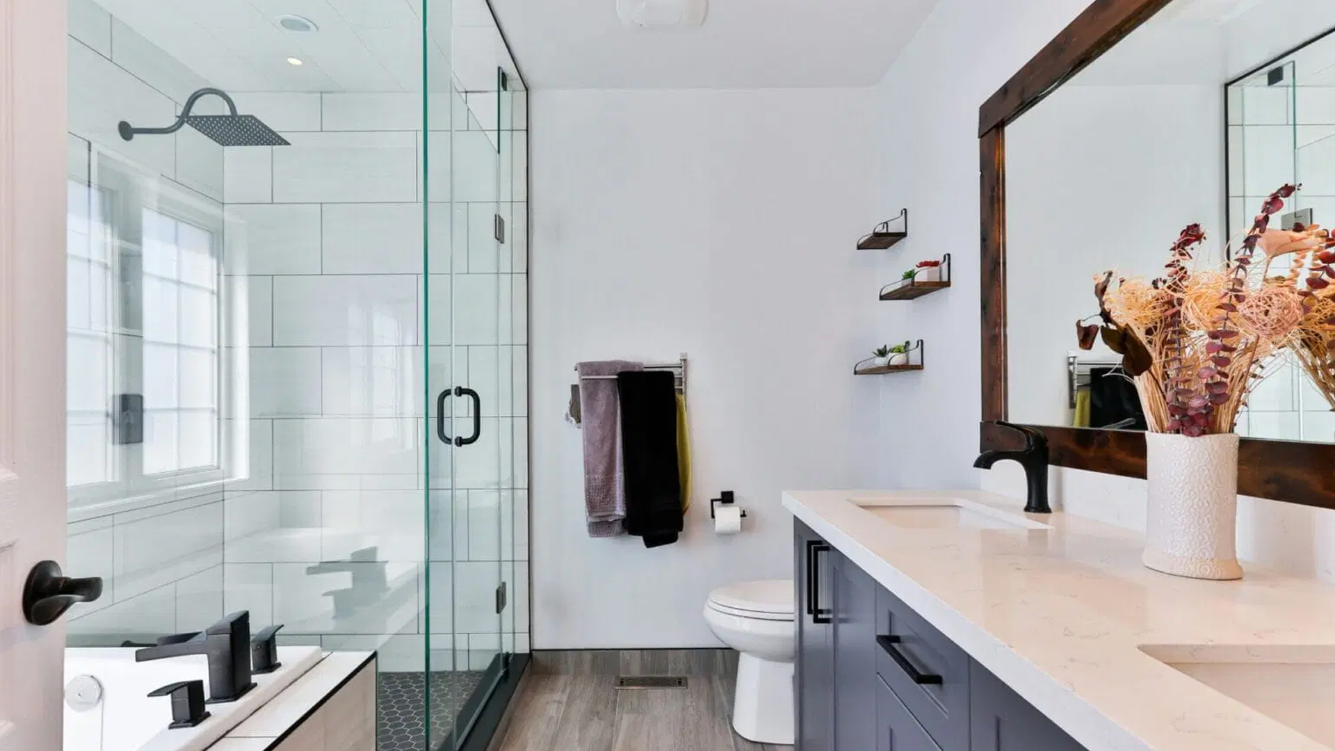 Modernes Badezimmer mit Glasdusche, weißem Waschbecken mit elegantem schwarzem Wasserhahn und Spiegel mit Holzrahmen. Regale, die mit Dekoration und Handtüchern verziert sind, schmücken die Wand. Neutrale Töne und minimalistisches Design zeigen die neuesten Trends bei der Badrenovierung.