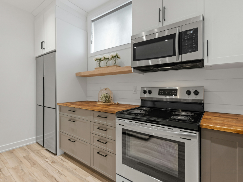Moderne Küche mit Edelstahlgeräten, Holzarbeitsplatten, weißen Schränken und einem kleinen Regal mit Pflanzen. Das Design fügt sich nahtlos in die angrenzenden Badmöbel ein.