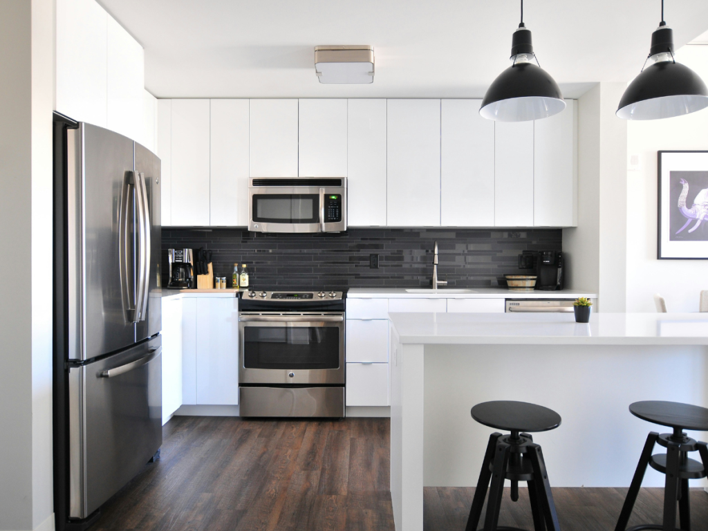Moderne Küche mit weißen Schränken, Edelstahlgeräten, einer dunklen Rückwand und zwei schwarzen Pendelleuchten über einer weißen Insel mit zwei schwarzen Hockern. Das schlanke Design spiegelt die Eleganz wider, die oft bei Badmöbeln zu finden ist.