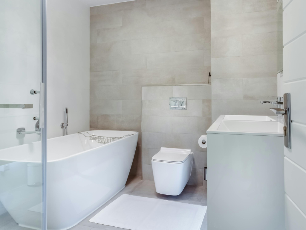 Moderne Badezimmerdesigns mit weißer Badewanne, wandmontierter Toilette und elegantem Waschbecken. Die beigen Fliesenwände sorgen für eine warme Note, ergänzt durch eine Duschtür aus Glas und eine kuschelige weiße Badematte auf dem Boden.