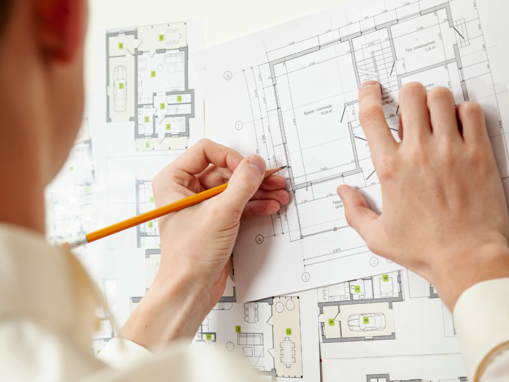 Eine Person mit einem Bleistift begutachtet sorgfältig den Entwurfsplan und die auf einem Tisch liegenden Architekturpläne.