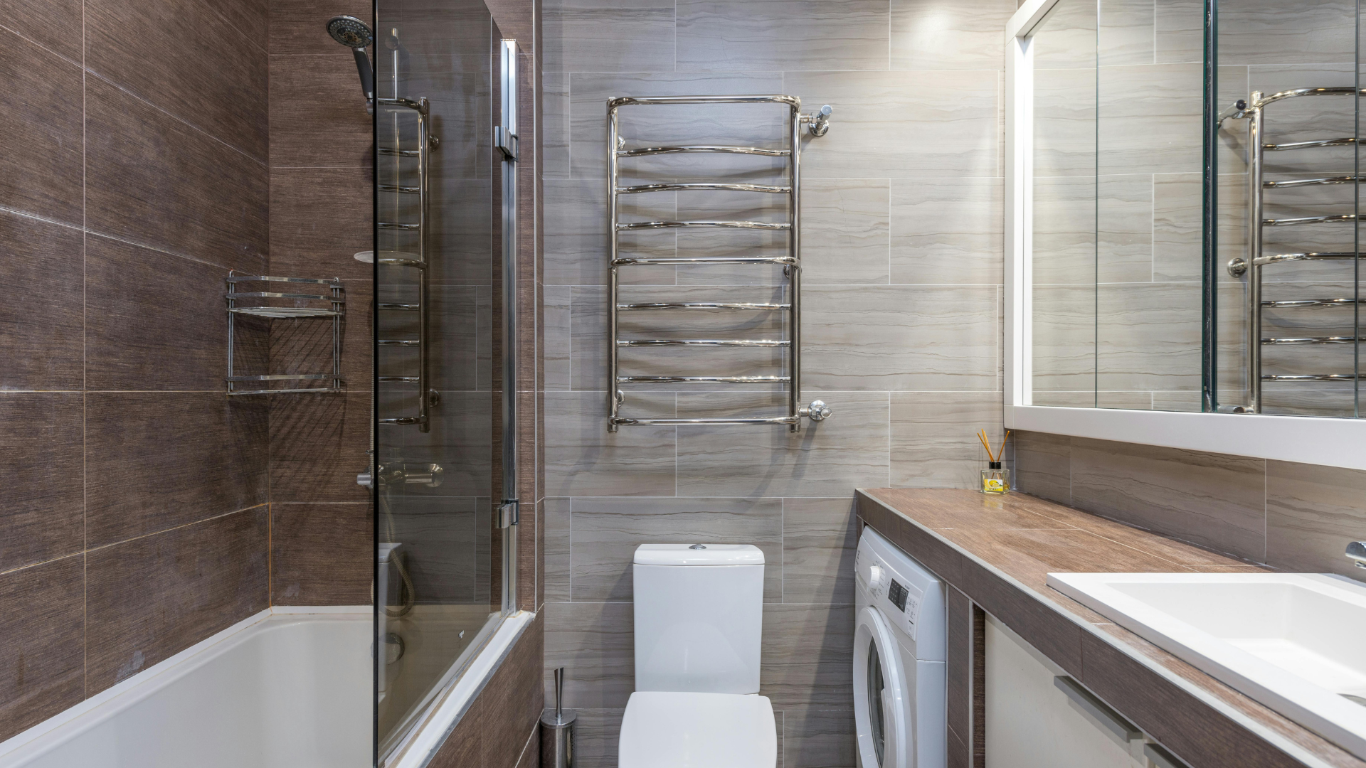 Ein modernes Badezimmer mit Badewanne, Glasduschtür, weißem WC und Handtuchwärmer erinnert an die behagliche Atmosphäre einer "warmen Küche". Es gibt eine Waschmaschine und ein Waschbecken mit Spiegel. Braune und beige Fliesen bedecken elegant die Wände und den Boden.