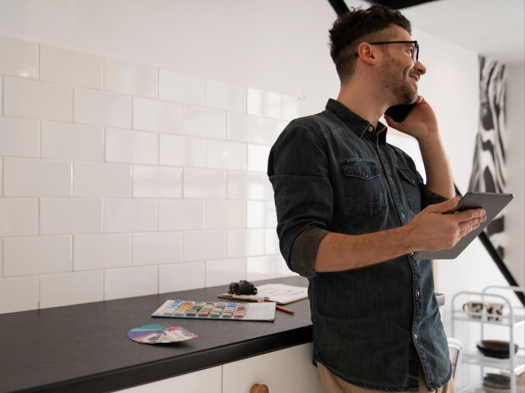 Ein Mann in Freizeitkleidung telefoniert, während er in einer Küche ein Tablet in der Hand hält. Küchengeräte sowie eine Farbpalette und eine Kamera sind auf der Arbeitsplatte verstreut.