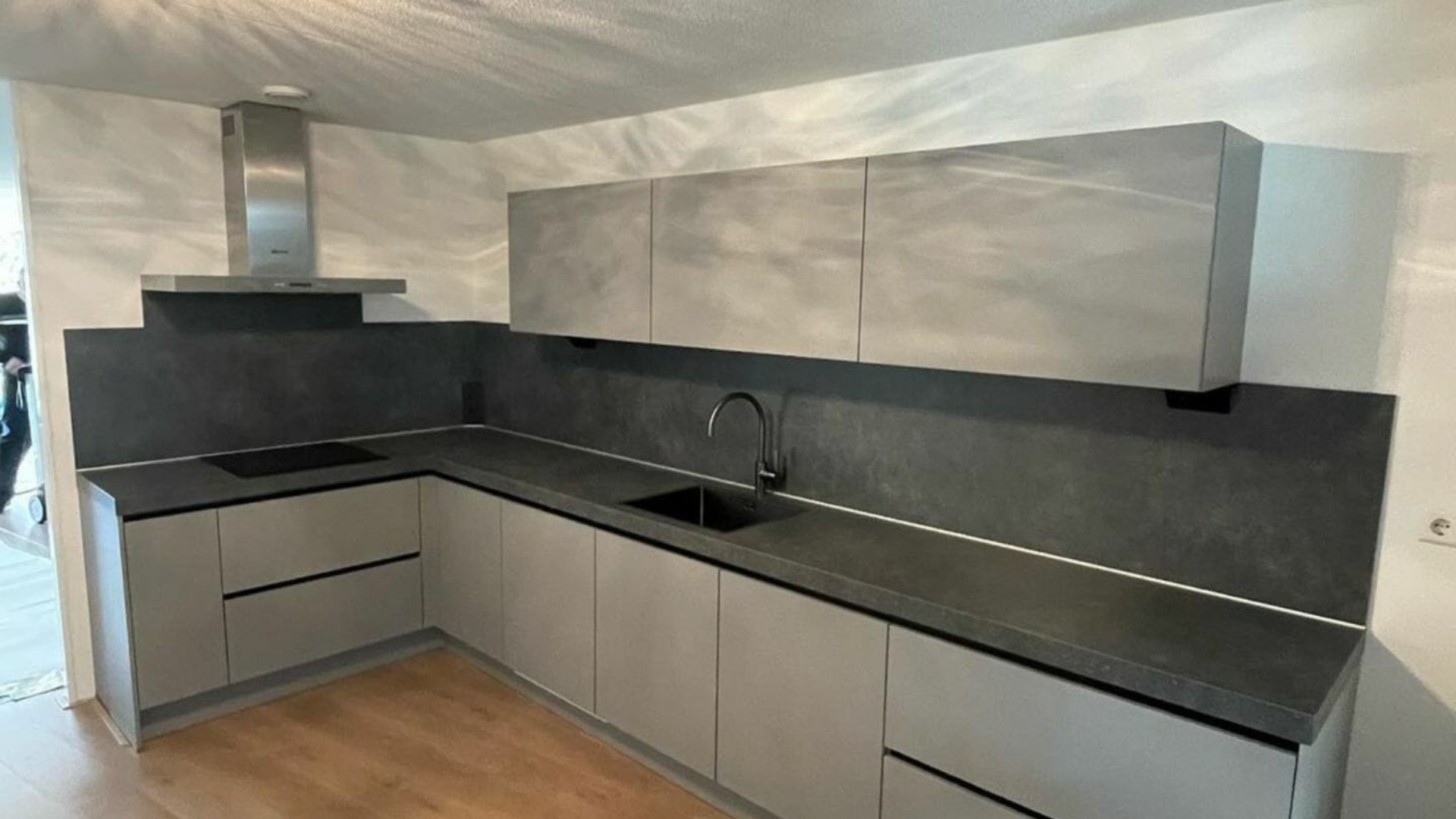 Moderne Küche mit mattgrauen Schränken, schwarzer Arbeitsplatte, eingebautem Kochfeld und Dunstabzugshaube aus Edelstahl. Helle Holzböden ergänzen das minimalistische Design und sind ein Paradebeispiel für Handwerkskunst in hochkarätigen Küchen- und Badprojekten.