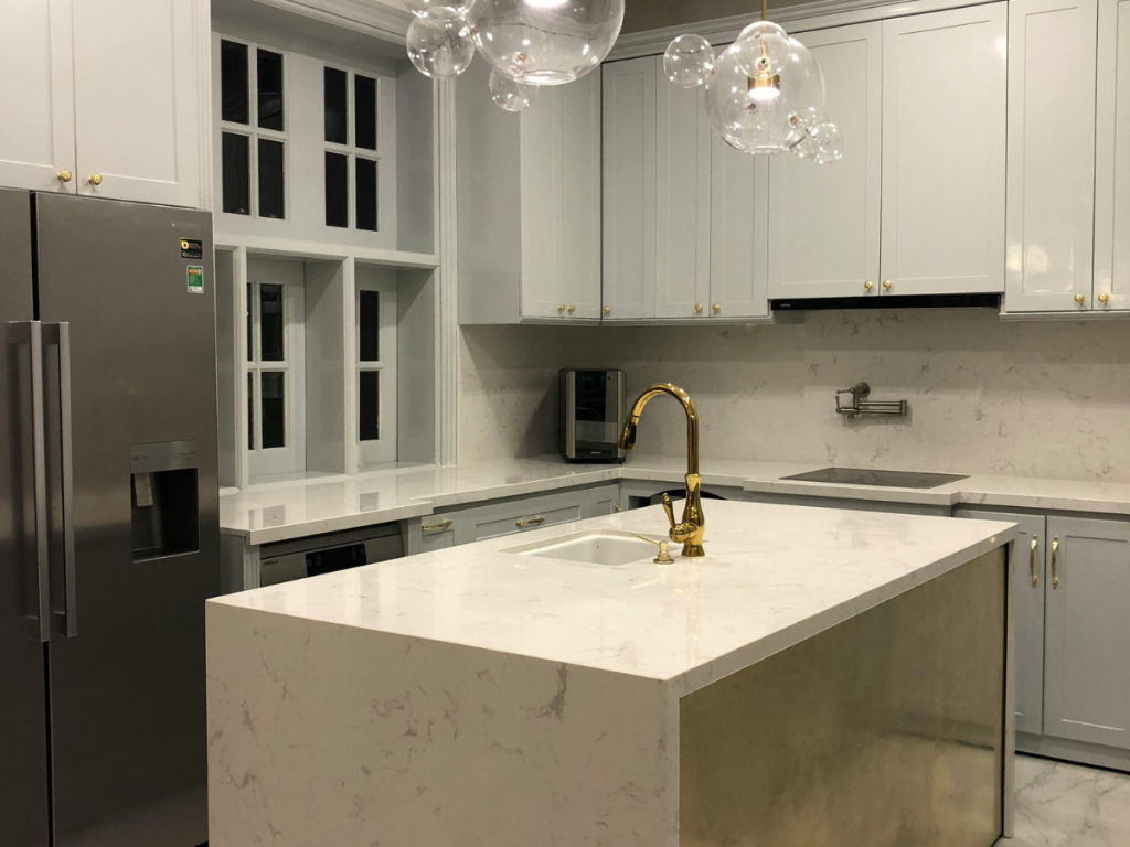 Moderne keuken met witte marmeren aanrechtbladen, een kookeiland met een gouden kraan en roestvrijstalen apparaten. Professionele begeleiding zorgt ervoor dat alles naadloos op elkaar is afgestemd. Hangende bubbellampen en strakke witte kasten maken de elegante ruimte compleet.