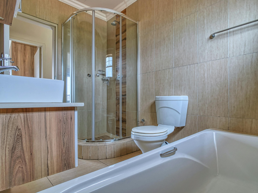 Ein modernes Badezimmer mit Glasdusche, weißem Waschbecken, WC und Badewanne. Die holzfarbenen Schränke und warmen Wandfliesen erinnern an die gemütliche Atmosphäre einer *warmen Küche* und fügen sich nahtlos in das schlichte Design ein.