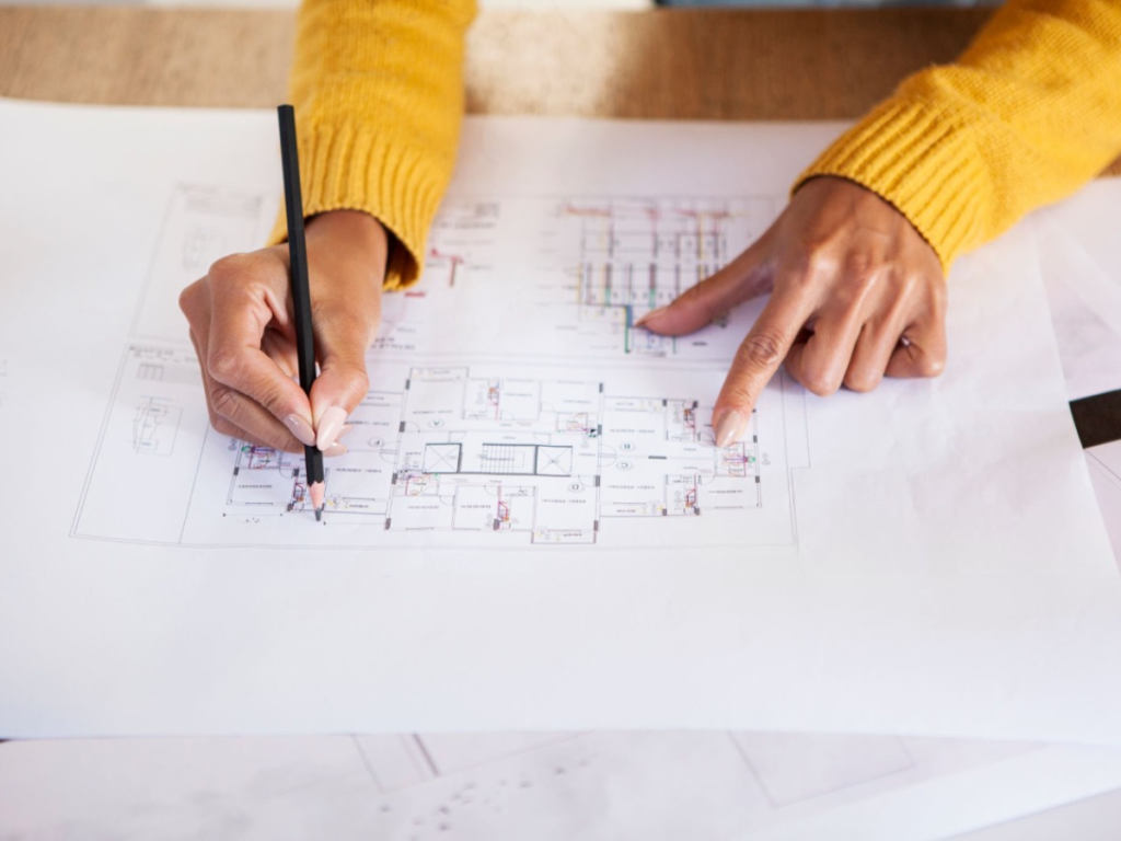 Eine Person im gelben Pullover zeichnet mit einem Bleistift präzise Architekturpläne und zeigt auf die filigranen Details ihrer Küchenentwürfe, die auf dem Tisch liegen.