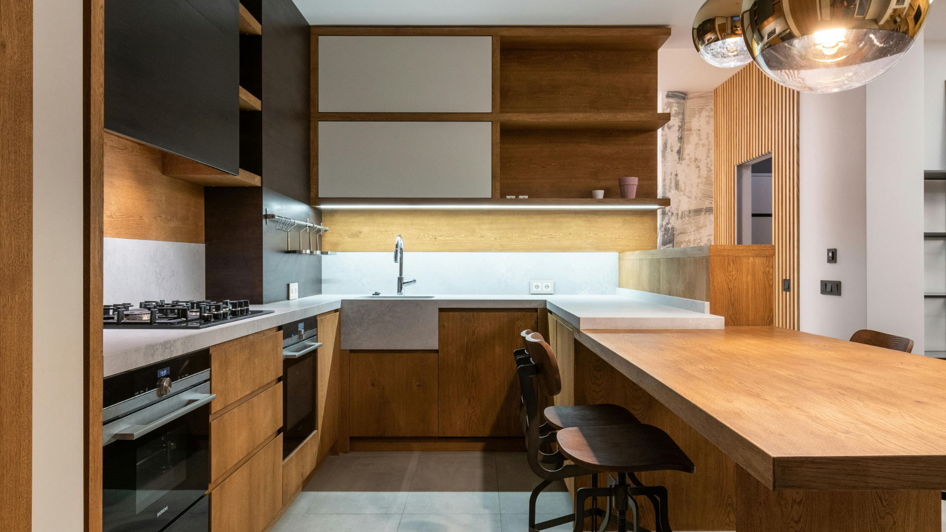 Die moderne Küche verfügt über elegante Holzschränke, eine elegante weiße Arbeitsplatte, einen Gasherd und einen Backofen. Im Vordergrund steht eine hölzerne Frühstückstheke mit Hockern unter stilvollen Pendelleuchten. Das Design fügt sich nahtlos in die Raffinesse ein, die oft in einer Luxusküche und einem luxuriösen Badezimmer zu finden ist.