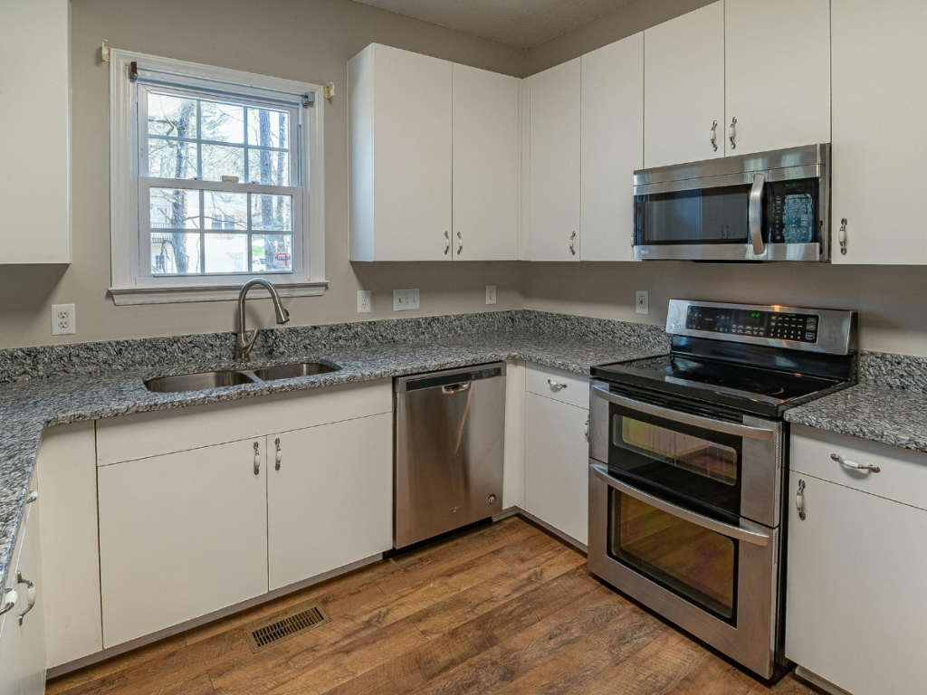 Moderne keuken met witte kasten, roestvrijstalen apparaten en granieten aanrechtbladen. Een dubbele spoelbak onder het raam biedt een perfect uitzicht, terwijl een lichte houten vloer de look compleet maakt. Geniet van professionele begeleiding bij het creëren van de keuken van uw dromen.