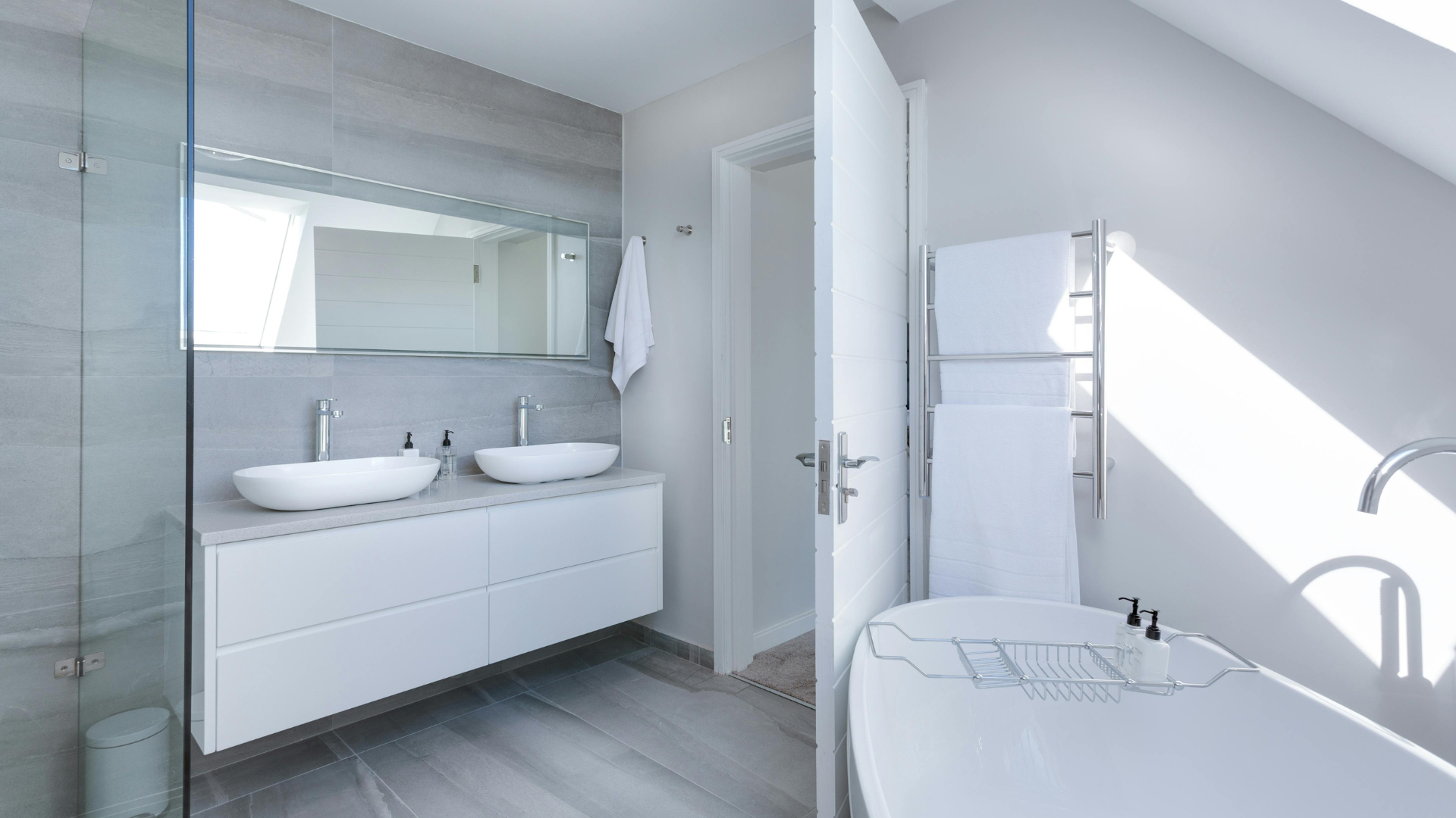 Modernes Badezimmer mit Doppelwaschbecken, großem Spiegel, freistehender Badewanne und Handtuchhalter. Hellgraue Fliesen und weiße Wände unterstreichen die geräumige, helle Atmosphäre, perfekt für alle, die sich für Badgestaltung interessieren.