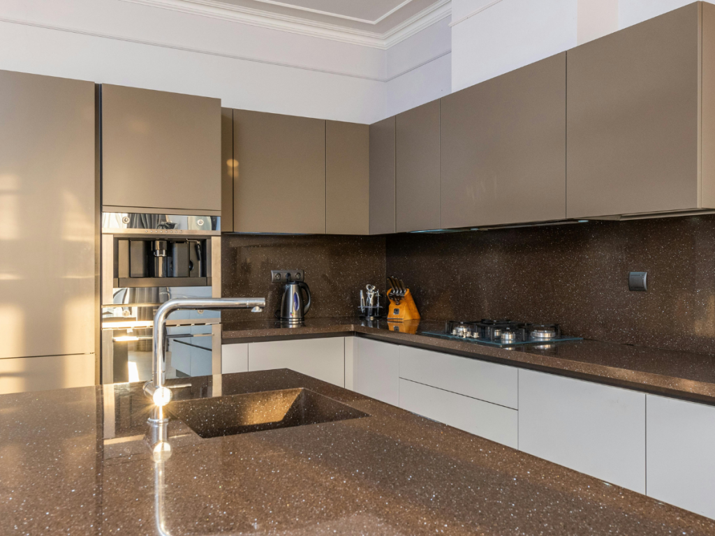Moderne Küche mit braunen Schränken, eingebautem Backofen und Kaffeemaschine aus Edelstahl. Braune Arbeitsplatte mit Spüle und Gasherd, gut beleuchtet durch natürliches Licht.