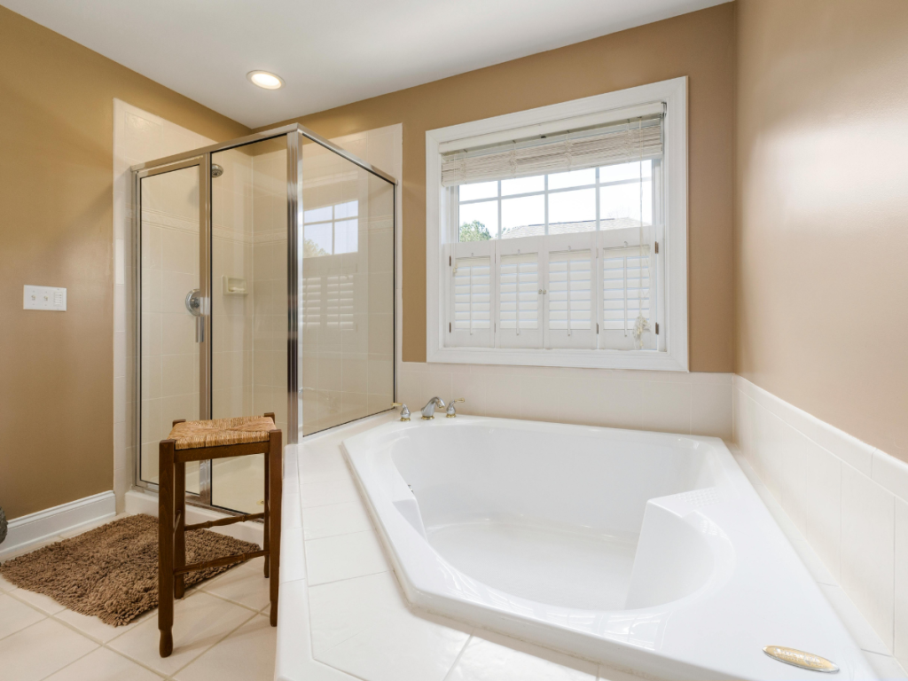 Ein Badezimmer mit einer großen weißen Badewanne, einer Glasdusche und einem kleinen Holzhocker verströmt die gemütliche Essenz einer warmen Küche. Das Sonnenlicht dringt sanft durch die Jalousien und verschönert dieses einladende Refugium.