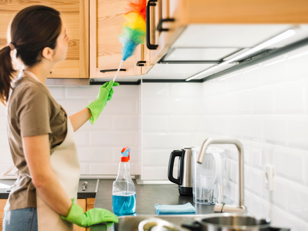 Eine Person staubt Küchenschränke mit einem Regenbogenstaubwedel ab, trägt grüne Handschuhe und eine Schürze. Ein Designplan bringt Symmetrie in die Szenerie, wobei eine Sprühflasche und ein Wasserkocher strategisch auf der Arbeitsplatte platziert sind.