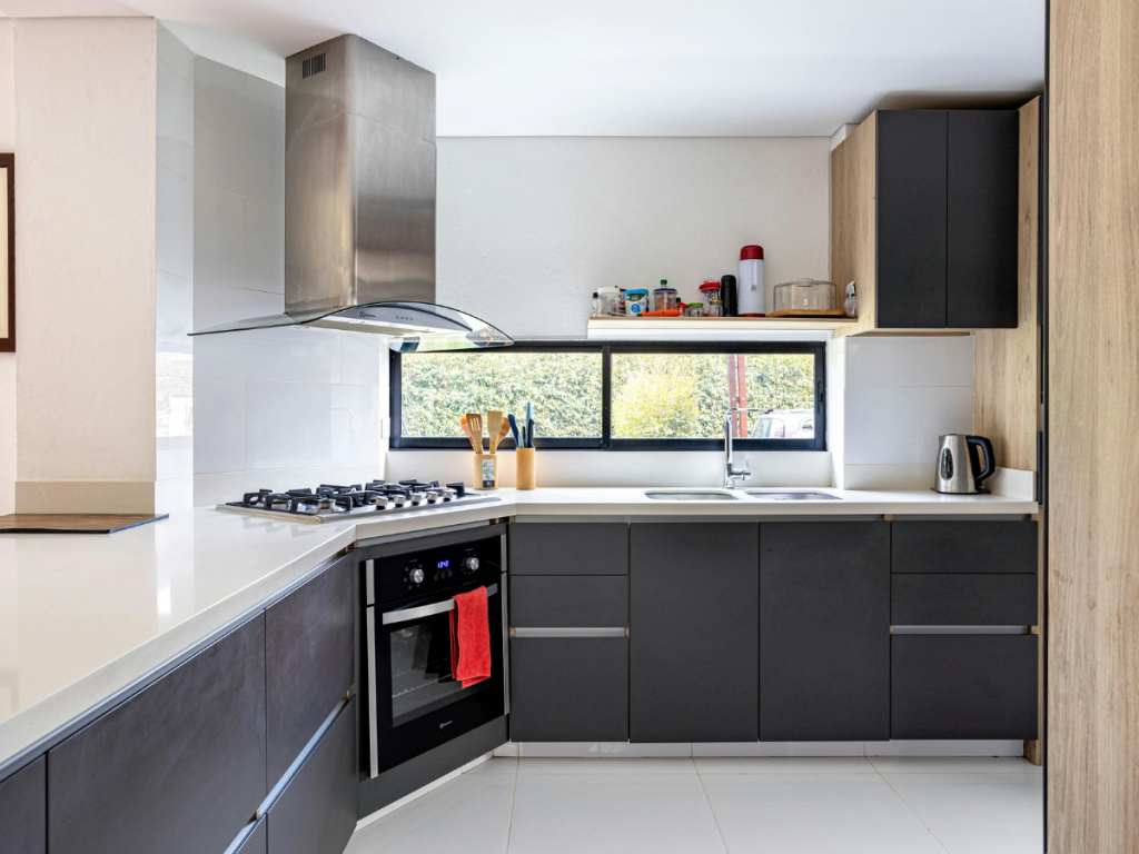 Moderne Küche mit eleganten schwarzen Schränken, einer Dunstabzugshaube aus Edelstahl und einem Gasherd. Ein großes Fenster über der Spüle lässt natürliches Licht herein und verbessert die Umgebung, in der die Geräte auf der Arbeitsplatte sichtbar sind. Dieser Raum bietet das Ambiente einer professionellen Anleitung für kulinarische Enthusiasten.