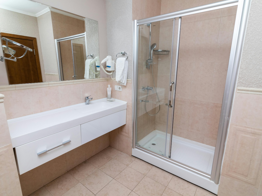 Ein modernes Badezimmer mit einer eleganten Glasdusche, einem weißen Waschbecken mit stilvollem Wasserhahn und großen Badezimmerspiegeln. Die beigefarbenen Fliesenwände und der Boden ergänzen das minimalistische Design, während Handtücher ordentlich auf einem Gestell in der Nähe des Waschbeckens hängen.