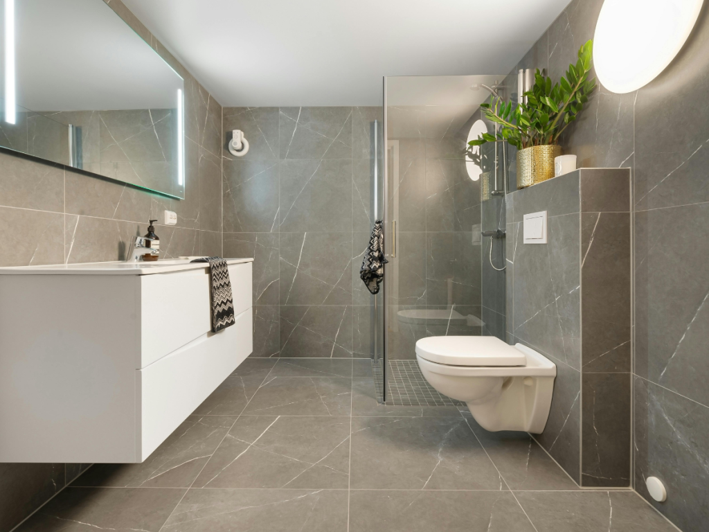 Ein modernes Badezimmer mit grauem Fliesenboden (Boden), einem weißen schwimmenden Waschbecken, einer wandhängenden Toilette und einer Glasdusche. Eine runde Lampe und Topfpflanzen runden die Einrichtung ab.