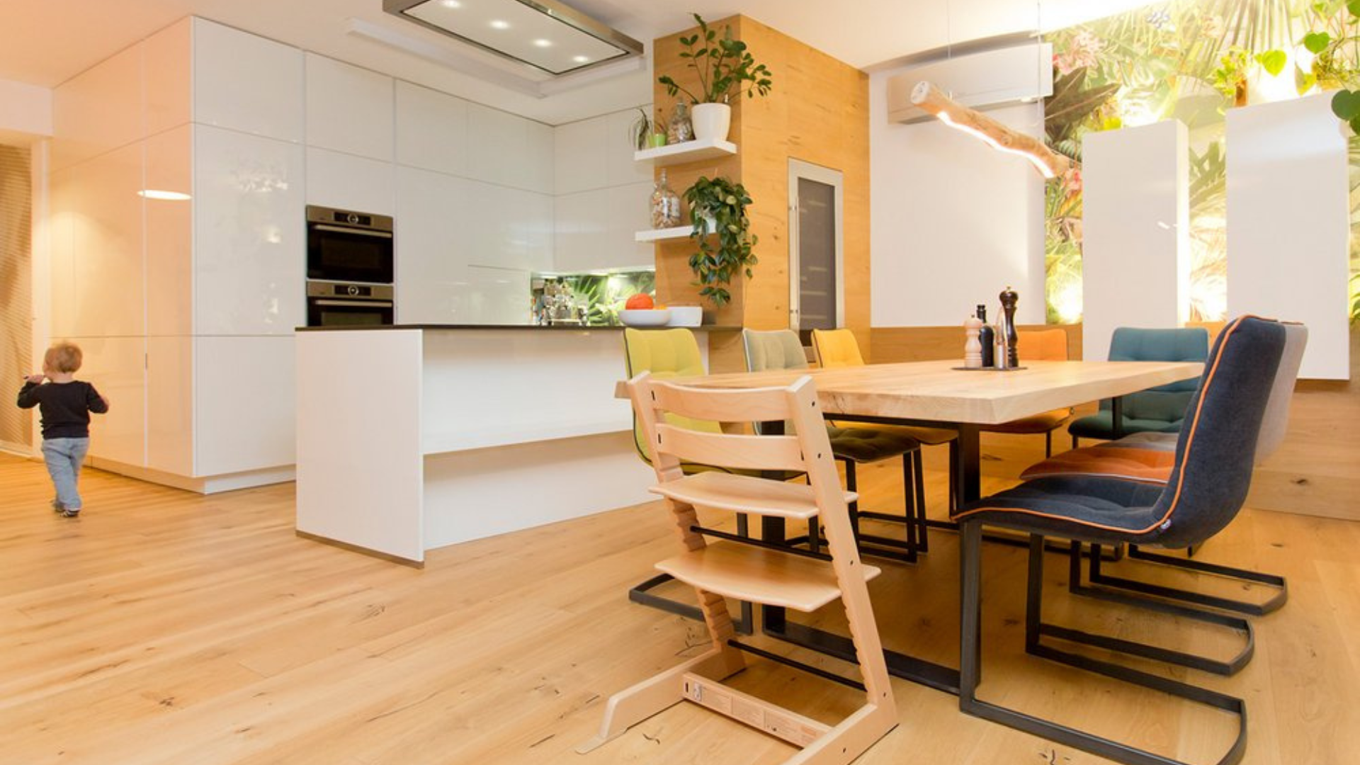 Eine moderne Küche und ein Essbereich mit weißen Schränken und bunten Stühlen um einen Holztisch sind der Inbegriff makelloser Inneneinrichtung, während ein Kind auf dem glatten Holzboden läuft.